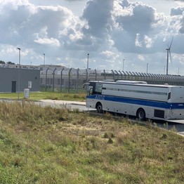 Ein blau-weißer Justiz-Bus fährt auf das Gelände der Forensik zu. Im Hintergrund ist der hohe Sicherheitszaun zu sehen. Der Himmel ist bewölkt.